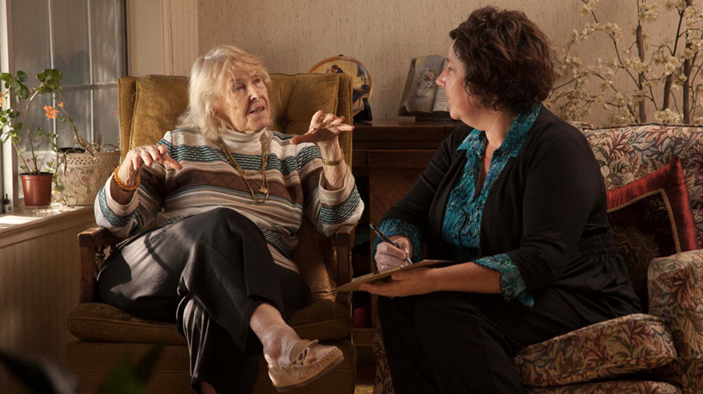 an image of a female social worker in business attire sitting with an elderly woman in her home chatting