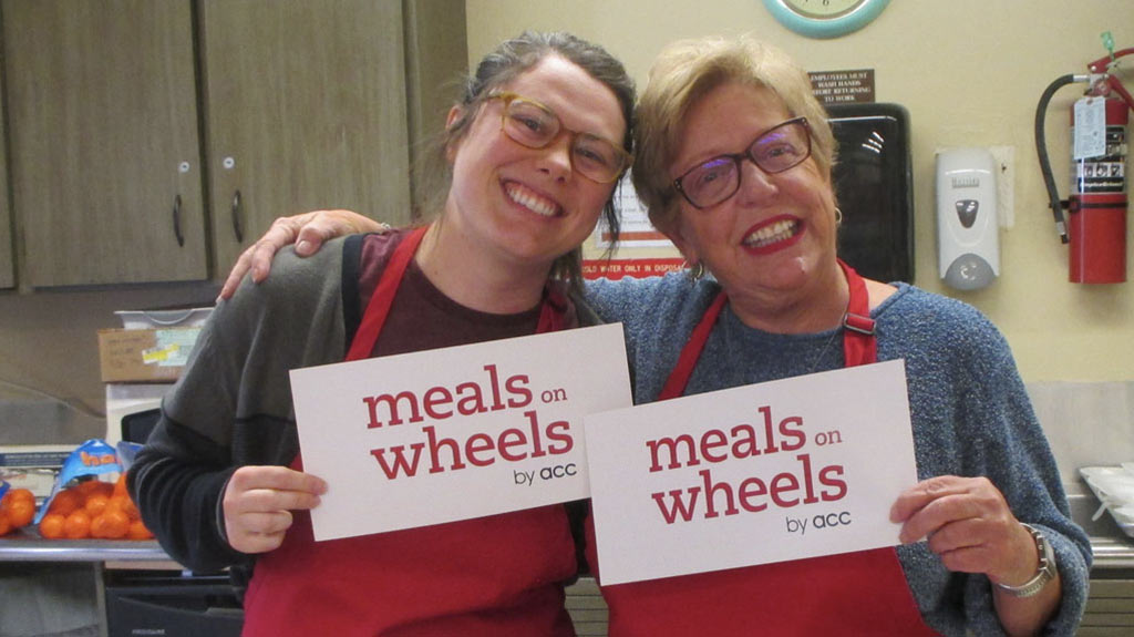 two women in red aprons standing together in the kitchen holding up signs that say Meals on Wheels by ACC
