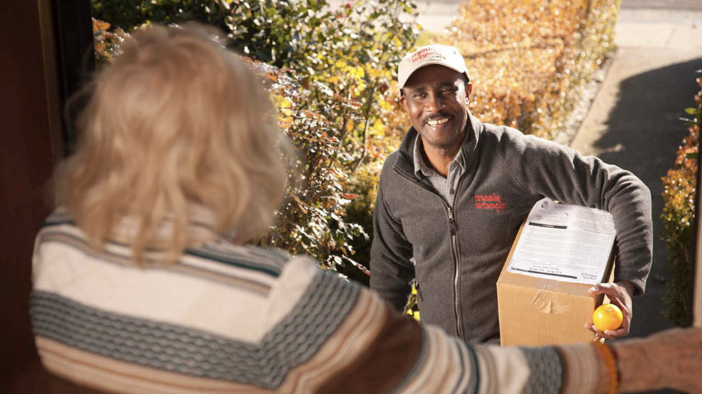 a meals on wheels delivery person delivering food to the front door of an elderly woman