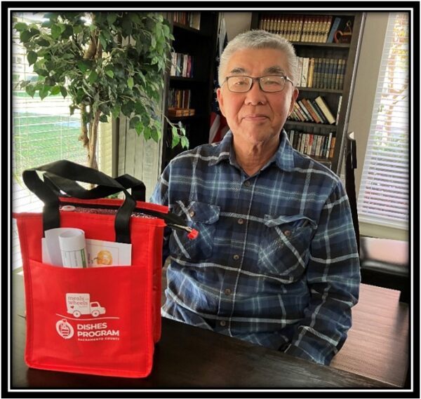 Volunteer Mr.Kim next to red bag of Meals on Wheels restaurant program.
